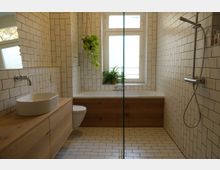 Modernes Badezimmer mit weiß gefliesten Wänden und Boden im Metrofliesen-Stil. Eine freistehende Badewanne mit Holzverkleidung steht vor einem Fenster, daneben eine Holzwaschtischplatte mit Aufsatzwaschbecken und einer Wandarmatur; an der Wand wachsen hängende Pflanzen.
