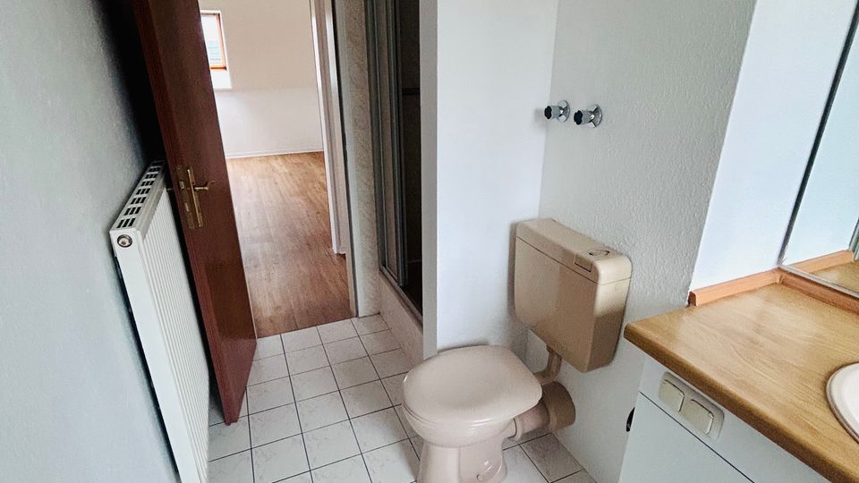 Schmales Badezimmer mit weiß gefliesten Bodenfliesen, WC und Waschtisch mit Holzplatte sowie Spiegelschrank. Links befindet sich ein Heizkörper und eine geöffnete Tür führt in einen angrenzenden Raum mit Holzfußboden.