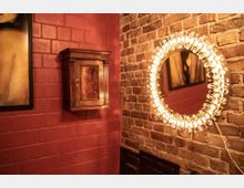 Ein Raum mit roten Ziegelwänden und einer rustikalen Holzwandkiste. Auf der rechten Seite ein beleuchteter runder Spiegel an einer unverputzten Backsteinwand, die den industriellen Stil des Raums unterstreicht.