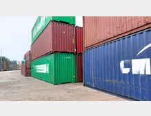 Stapel von mehrfarbigen Schiffscontainern (grün, rot und blau) auf einem großen, asphaltierten Lagergelände mit wenigen Bäumen im Hintergrund. Die Container sind in mehreren Ebenen übereinandergestapelt.