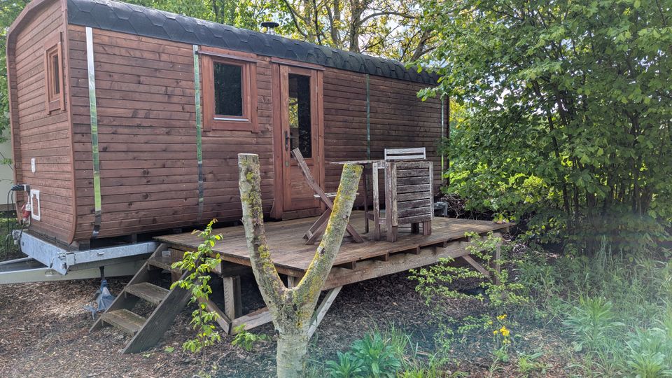 Holzhütte auf einem Anhänger mit schwarzem Schindeldach, Fenster und Tür, davor eine erhöhte Holzterrasse mit Stufen und einfachen Holzmöbeln. Die Hütte steht in einer grünen, von Bäumen und Sträuchern umgebenen Garten- oder Waldrandumgebung.