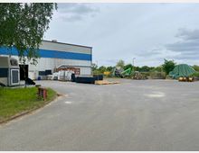 Industrieller Hofbereich mit einer großen Wellblechhalle, gestapelten Materialien und Maschinen. Im Vordergrund eine offene Asphaltfläche, im Hintergrund Bäume und ein eingepackter Materialhaufen.