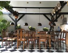 Ein rustikales Esszimmer mit einem langen Holztisch und verschiedenen, antik wirkenden Stühlen vor einer Wand mit schwarzen Holzbalken. Über dem Tisch hängen drei schwarze Pendelleuchten, und auf dem Tisch stehen dekorative Blumenarrangements; der Boden ist im Schachbrettmuster gefliest.