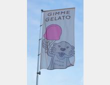 Flagge mit der Aufschrift „Gimme Gelato“ und einer gezeichneten Otterfigur, die ein rosafarbenes Eis hält. Darunter steht „Berlin Est. 2018“; der Hintergrund zeigt einen blauen Himmel.