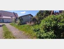 Wohngrundstück mit einem kleinen Haus mit rotem Ziegeldach und einem Garten, der von dichtem Grün und Büschen umgeben ist. Ein gepflasterter Weg führt durch den Garten, der teils von Gras und Pflanzen überwuchert ist.