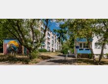 Wohngebiet mit mehrstöckigen Wohnhäusern und Grünflächen. Links ein kleines Gebäude mit einer bunten Wandbemalung, auf der ein Eisvogel abgebildet ist, rechts Bäume und ein Gehweg.