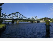 Grüne Stahlbrücke mit Fachwerkstruktur, die über einen breiten Fluss führt, umgeben von grünen Bäumen und einer kleinen steinernen Uferbebauung. Der Himmel ist klar und blau.