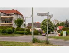 Wohngebiet mit Einfamilienhäusern und mehrstöckigen Gebäuden im Hintergrund. Im Vordergrund eine Straßenkreuzung mit Straßenschildern, einem Laternenmast und gepflegten Gärten, umgeben von Bäumen und Sträuchern.