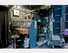 Ansicht eines Technikraums hinter der Bühne in einem Theater oder Veranstaltungsort. Zu sehen sind ein Mischpult mit Monitoren, zahlreiche Kabel, Schalter und technische Geräte, sowie ein schwarzer Vorhang und eine große Wanduhr.