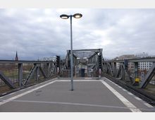 Eine erhöhte Bahnstation mit Schienen, die über eine stählerne Gitterbrücke führen. Im Hintergrund sind moderne Wohngebäude und die Spitze einer Kirche zu sehen, bei bewölktem Himmel.
