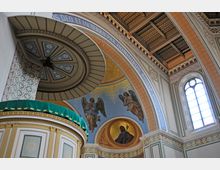 Innenraum einer Kirche mit detaillierten Wand- und Deckengemälden. Zu sehen sind ein geschmückter Kanzelaufbau mit grünem Stoffbaldachin, ein goldener Bogen mit lateinischer Inschrift, Fresken von Engelmotiven und eine runde Fensterglasöffnung mit Lichtschein.