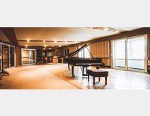 Ein modernes Musikstudio mit einem Flügel im Mittelpunkt, umgeben von Akustikplatten an den Wänden und Fenstern mit Tageslichteinfall. Im Hintergrund stehen Musikinstrumente, Verstärker und Regale, die den Raum funktional gestalten.