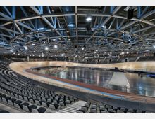 Innenansicht einer großen, modernen Radrennbahn-Halle mit ovaler Holzbahn und großem Innenfeld, umgeben von abgestuften Tribünen mit schwarzen Sitzreihen. Über der Arena spannt sich eine weitläufige Stahlträger-Decke mit integrierter Beleuchtung und hängender Veranstaltungstechnik.