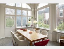 Esszimmer in einem hellen Wintergarten mit großen, bogenförmigen Fenstern; ein Holzesstisch ist umgeben von gepolsterten Stühlen, mit Schalen voller Äpfel darauf, und die Fensterfront bietet Blick auf einen Garten sowie Backsteingebäude im Hintergrund.