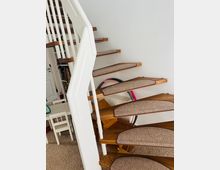 Eine moderne Wendeltreppe aus Holz mit Teppichläufern auf den Stufen und einem weißen Geländer. Im Hintergrund sind Möbel und Gegenstände wie ein Kinderstuhl, ein Tisch und bunte Hula-Hoop-Reifen zu sehen.