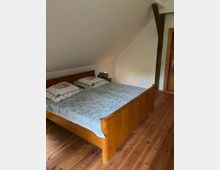 Schlafzimmer mit Holzboden und einem doppelten Holzbett mit gemusterten Bettbezügen. Der Raum hat eine schräge Decke, eine sichtbare Holzbalkenkonstruktion und eine Tür mit Holzrahmen.