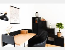 Minimalistisch eingerichtetes Arbeitszimmer mit einem Holztisch, einem schwarzen Stuhl und schwarzen Regalmöbeln. An der weißen Wand hängt eine Karte von Berlin, und dekorative Elemente wie Pflanzen, Bücher und eine Lampe ergänzen den Raum.