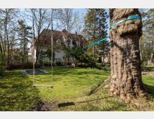 Garten mit Baum, an dem eine Slackline befestigt ist, sowie eine Schaukel, die zwischen dem Baum und einem weiteren Punkt hängt. Im Hintergrund steht ein Haus mit rotem Ziegeldach und weißen Wänden, umgeben von Bäumen und Sträuchern.