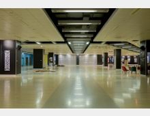 Weitläufiger, moderner Innenraum mit hell glänzendem Bodenbelag, mehreren schwarzen Säulen und abgehängten Decken mit integrierter Beleuchtung. Im Hintergrund befinden sich geschlossene Rolltore, bunte Wände und Sitzbereiche mit bunten Stühlen.