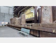 Industriehalle mit einer massiven Anlage aus rostigem Metall, bestehend aus großen Säulen und Platten mit sichtbarer Nummerierung. Im Vordergrund steht eine hellblaue Metallbank, während im Hintergrund Wände aus Beton und ein verglastes Bürogebäude zu sehen sind.