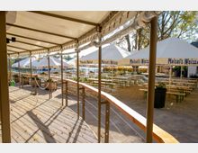 Sonniger Biergarten mit Holzbänken und Tischen unter großen weißen Sonnenschirmen mit der Aufschrift „Maisel’s Weisse“. Im Vordergrund ein überdachter Holzsteg mit Geländer, im Hintergrund Bäume und eine Terrasse.