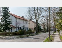 Straßenszene in einem Wohngebiet mit zweigeschossigen Einfamilienhäusern, weißen und beigen Fassaden und roten Ziegeldächern. Die Straße ist gesäumt von Bäumen und Gärten, mit einem Gehweg auf beiden Seiten sowie Verkehrsschildern, die ein Tempolimit von 50 km/h anzeigen.