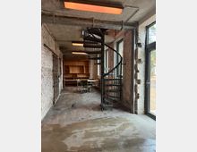Industrieller Raum mit Betonboden, rauen Ziegelwänden und einer Wendeltreppe aus Metall. Im Hintergrund stehen ein Holzkastenregal und ein Tisch mit Stühlen, flankiert von großen Fenstern mit Blick nach außen.