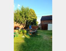 Grüner Außenbereich mit mehreren Holztischen und -stühlen auf einer Rasenfläche, umgeben von Bäumen, einer Backsteinmauer und einem Gartenschuppen mit Holztüren. Lichterketten sind in der Luft gespannt, und der Bereich ist in warmem Sonnenlicht beleuchtet.