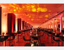 Großer Veranstaltungsraum mit langen, weißen Säulen, dunklen Tischen und Stühlen sowie einer stimmungsvollen Beleuchtung in warmen Rot- und Gelbtönen. Die Tische sind mit Gläsern, Kerzen und Blumen dekoriert, und rote Vorhänge säumen die Wände.