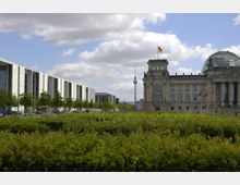 Ansicht des Reichstagsgebäudes in Berlin mit seiner gläsernen Kuppel und der deutschen Flagge auf dem Dach. Im Vordergrund sind gepflegte Grünanlagen und Büsche, links daneben verläuft ein modernes Bürogebäude, und im Hintergrund ist der Berliner Fernsehturm zu sehen.
