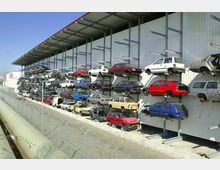 Mehrstöckige Lagerung von Autos an einer Metallfassade eines großen Gebäudes, vermutlich ein Schrottplatz oder eine Autoverwertung. Im Vordergrund befindet sich ein Zaun mit Stacheldraht.