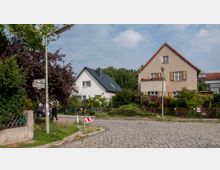 Straßenecke in einem Wohngebiet mit Kopfsteinpflaster, einem Straßenschild "Königstuhlweg" und zwei freistehenden Einfamilienhäusern mit Gärten, eines weiß mit schwarzem Dach und eines beige mit Holzläden und Blumen im Fenster. Im Hintergrund sind Bäume und weitere Häuser zu sehen.