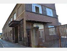 Zweistöckiges Gebäude mit teils verputzter und teils verklinkerter Fassade, großen Fenstern und einem überdachten Eingang. Im Vordergrund ein verwitterter Metallzaun und eine schmale, gepflasterte Gasse.