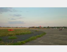 Weitläufiges ehemaliges Flughafenareal mit Betonflächen und Grünstreifen, im Hintergrund eine Stadtansicht mit Gebäuden. Ein Fahrradfahrer mit Hund ist auf der rechten Seite sichtbar, zudem ein Runway-Schilderpaar mit der Aufschrift "09L" und "09R" im Vordergrund.