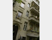 Alt-Text: Mehrstöckiges Wohngebäude im Altbaustil mit verzierten Fassaden, Balkonen und großen Fenstern. Vor dem Haus steht ein Fahrrad, und einige Balkone sind mit Pflanzen dekoriert.