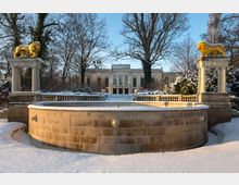 Historisches Herrenhaus in einem verschneiten Park, im Vordergrund ein steinernes Brunnenbecken mit Löwenmotiven und zwei goldenen Löwenstatuen auf Säulen. Die Umgebung ist von kahlen Bäumen und Sträuchern umgeben, beleuchtet von tiefstehendem Sonnenlicht.