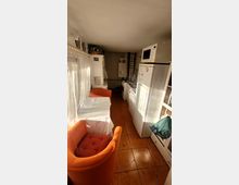 Schmaler Küchen- und Wohnraum mit orangefarbenen Sesseln und einem kleinen Tisch mit weißer Tischdecke auf der linken Seite. Auf der rechten Seite befinden sich ein Kühlschrank, eine Mikrowelle und andere Küchengeräte, der Boden ist mit quadratischen, braunen Fliesen ausgelegt.