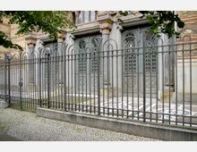 Fassade eines historischen Gebäudes mit verzierten Metalltüren und steinernen Säulen, umgeben von einem hohen, dekorativen Metallzaun. Im Vordergrund befindet sich ein gepflasterter Gehweg.