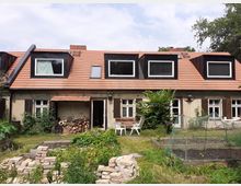 Rückansicht eines niedrigen, länglichen Wohnhauses mit rotem Ziegeldach und mehreren dunklen Gaubenfenstern sowie Holzläden an den Fenstern. Davor liegt ein Garten mit Hochbeeten, Rasen, gestapelten Ziegeln und einem kleinen Sitzbereich neben einem überdachten Holzlager.