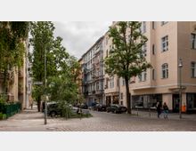 Straßenszene in einer Wohngegend mit mehrstöckigen Altbauten, Bäumen und Kopfsteinpflaster. Ein Café befindet sich im Erdgeschoss eines Gebäudes, parkende Autos stehen entlang der Straße, und Passanten sind auf dem Gehweg zu sehen.