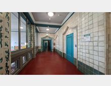 Langer Innenflur in einem Gebäude mit rotbraunem Boden, weiß gekachelten Wänden und runden Deckenleuchten. Rechts befindet sich eine türkisfarbene Tür, links große Fenster und dekorierte Türrahmen mit blaugrünen Ornamenten.