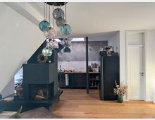 Moderne Wohnküche mit einem offenen Kamin in schwarzem Design, einem schwarzen Kühlschrank und einer grauen Küchenzeile mit weißen Fliesen. Eine Lampe mit mehreren gläsernen Kugeln hängt von der Decke, und auf dem Holzboden steht eine Vase mit Trockenblumen.