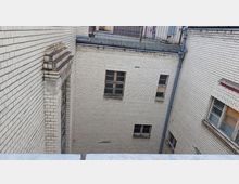 Innenhof eines Gebäudes mit hellen Ziegelwänden. Mehrere kleine Fenster mit teils beschädigten oder zugemauerten Holzrahmen sind sichtbar, umgeben von Regenrinnen und einem niedrigen Metallgitter auf dem Dachrand.