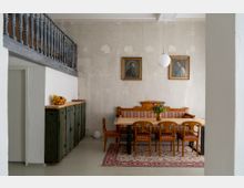 Esszimmer mit rustikalem Holztisch, sechs gepolsterten Stühlen und einer Holzbank vor einer Wand mit zwei gerahmten Porträts. Ein Teppich liegt unter dem Tisch, und eine Hängelampe beleuchtet den Raum. Im Hintergrund steht ein grünes Schrankmöbel mit Obstschale.