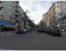 Straßenszene in einer Stadt mit mehrstöckigen Wohn- und Geschäftsgebäuden zu beiden Seiten, geparkten Autos am Straßenrand und Geschäften im Erdgeschoss. Fußgänger und ein Radfahrer sind auf der Straße zu sehen, während kahle Bäume die Straße säumen.