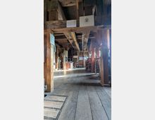 Innenraum eines historischen Holzgebäudes mit freiliegenden Deckenbalken, breiten Holzdielen und mehreren Stützpfosten. Entlang der Wände stehen alte Maschinen und Ausstellungsstücke, ergänzt durch Informationstafeln; Tageslicht fällt durch seitliche Fenster ein.