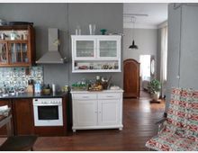 Eine offene Küche in einem Wohnraum mit Holzboden; links befindet sich eine Küchenzeile mit dunklen Holzschränken, Herd und Dunstabzugshaube sowie bunt gemusterten Fliesen. Rechts steht ein weißer Küchenschrank, im Hintergrund sind ein Holzschrank und ein kleiner Tisch mit Pflanzen zu sehen.