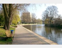 Eine gepflasterte Uferpromenade an einem Fluss mit Bäumen am Rand, einer Parkbank und einem Papierkorb im Vordergrund; im Hintergrund sind ein moderner Gebäudeblock und weitere Bäume zu sehen.