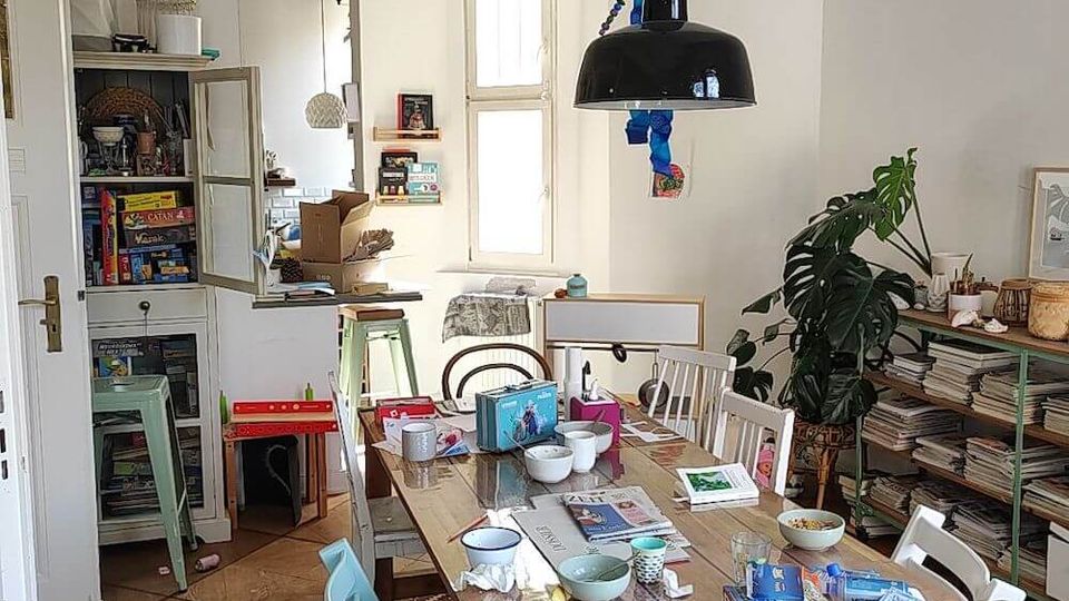 A dining room or multi-purpose space with a wooden table cluttered with dishes, books, and papers, surrounded by mismatched chairs. The room features white walls, wooden flooring, a large hanging black light fixture, tall windows, shelves filled with magazines, and a large potted plant.