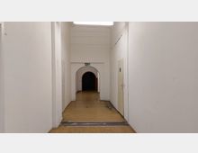 Ein langer, schlichter Innenflur mit weißen Wänden und einem Boden aus hellem Holzimitat. Am Ende des Flurs befindet sich ein Rundbogen mit einem grünen Fluchtwegschild darüber, der in einen dunkleren Raum führt; rechts im Flur ist eine geschlossene Tür sichtbar.